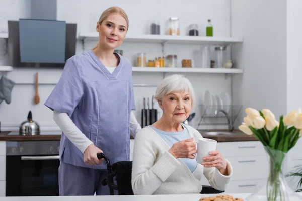 年轻的护士看着镜头靠近残疾的老妇人 在厨房里端着一杯茶 — 图库照片