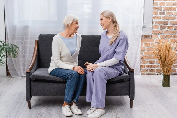 Jovem Assistente Social Mulher Idosa Sorrindo Enquanto Conversa Sofá Casa — Fotografia de Stock