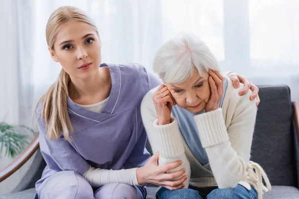 Young Nurse Looking Camera While Embracing Senior Woman Sitting Bowed — Stock Photo, Image