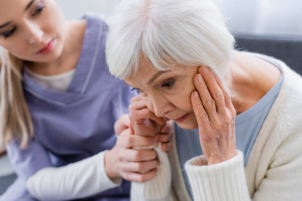 young nurse touching hand of elderly woman, suffering from dementia and sitting with bowed head, blurred background