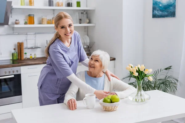 Młoda Pielęgniarka Uśmiecha Się Kamery Obejmując Szczęśliwą Starą Kobietę Kuchni — Zdjęcie stockowe