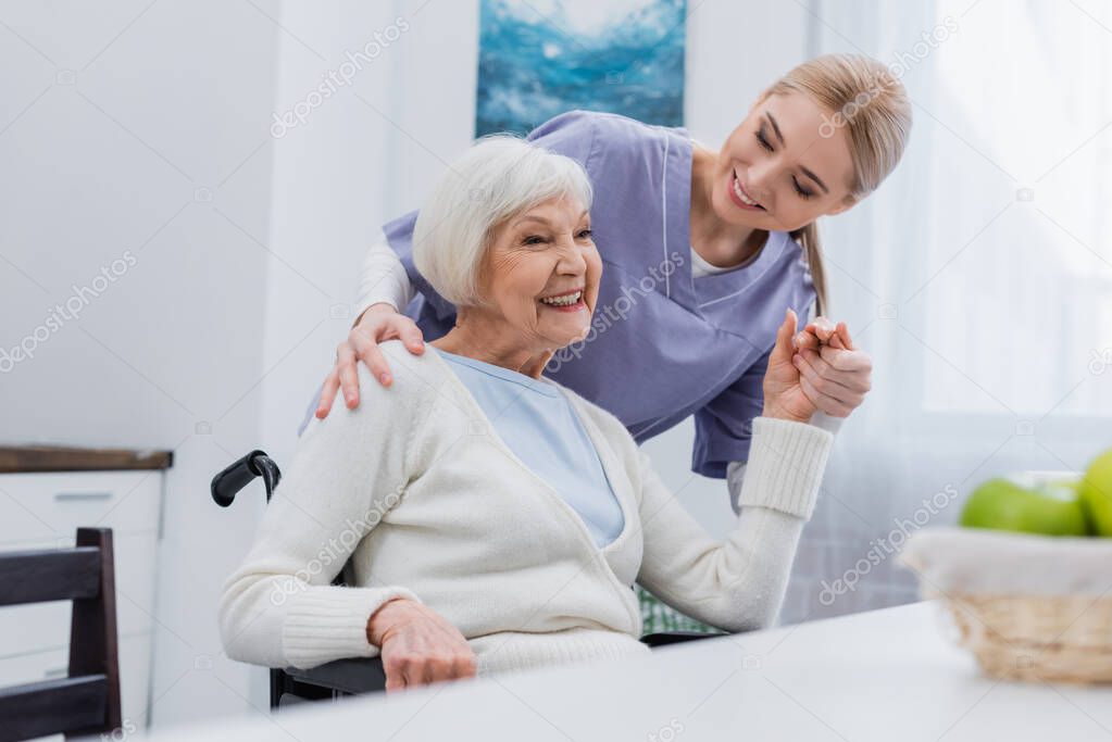 smiling nurse holding hands with joyful handicapped woman at home