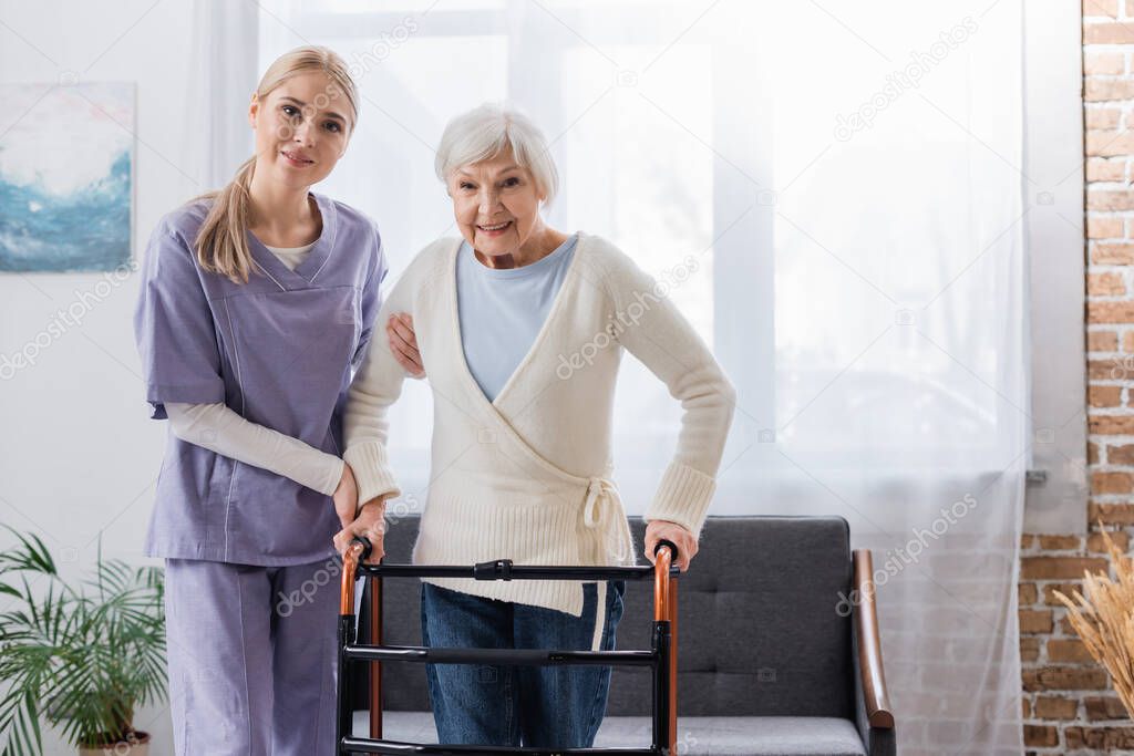 happy nurse and senior woman looking at camera near medical walkers in living room