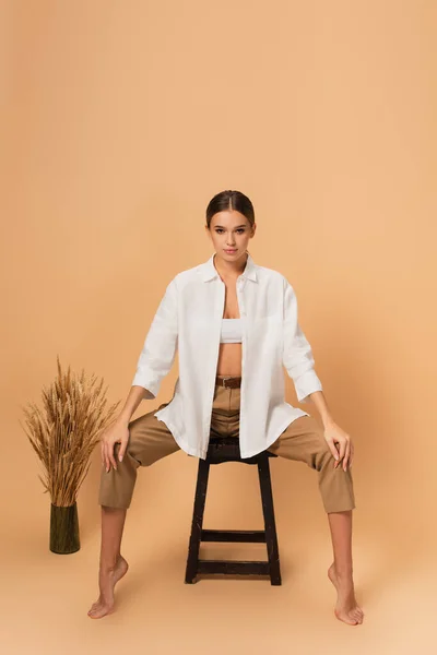 barefoot woman in pants and unbuttoned shirt sitting on wooden chair on beige background