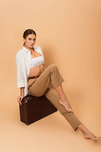 sensual barefoot woman in pants and unbuttoned shirt posing on vintage suitcase on beige