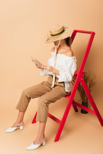 young woman in straw hat, white shirt and pants reading book on ladder near spikelets on beige 