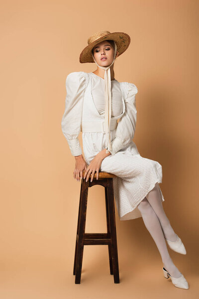 В полный рост молодая женщина в белом платье и соломенной шляпе сидит на табуретке бежевого цвета