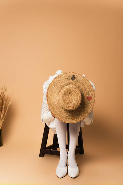 young woman in straw hat obscuring face and sitting on wooden chair near wheat spikelets on beige