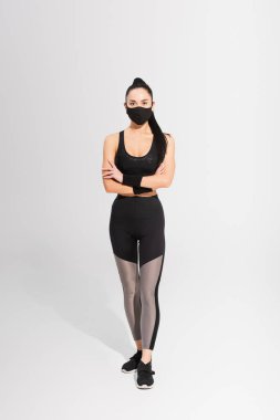young sportswoman in sportswear and black protective mask standing with crossed arms on grey clipart