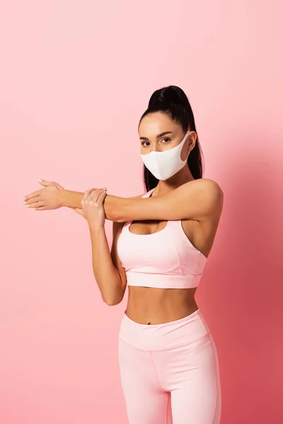 Ung Sportskvinne Med Medisinsk Maske Som Strekker Armen Mot Rosa – stockfoto