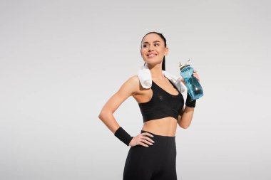 Mutlu sporcu kadın omuzlarında havluyla spor şişesi tutarken elleri kalçasında gri renkte poz veriyor.