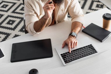 Kesilen adamın grafik tablet, defter ve kağıt bardak yanında dizüstü bilgisayar kullanmasının yüksek açılı görüntüsü