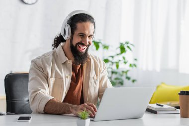 Evde çalışırken dizüstü bilgisayarda yazan kulaklıkla gülen İspanyol serbest yazar.