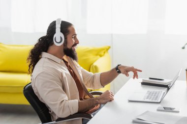 Çevrimiçi Webinar sırasında dizüstü bilgisayarı işaret eden kulaklıklı neşeli İspanyol serbest yazar