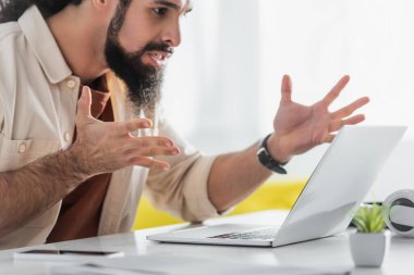 irritated hispanic freelancer gesturing near computer at home clipart