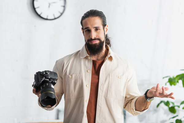 latin man with digital camera showing shrug gesture while standing in home studio