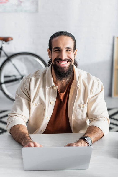 счастливый латиноамериканец смотрит в камеру, работая дома на ноутбуке