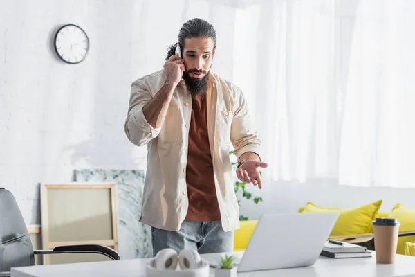 Hombre Hispano Hablando Teléfono Inteligente Apuntando Computadora Portátil Escritorio Casa — Foto de Stock