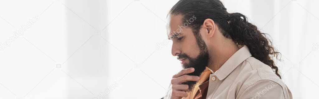 side view of pensive hispanic man touching beard while thinking at home, banner