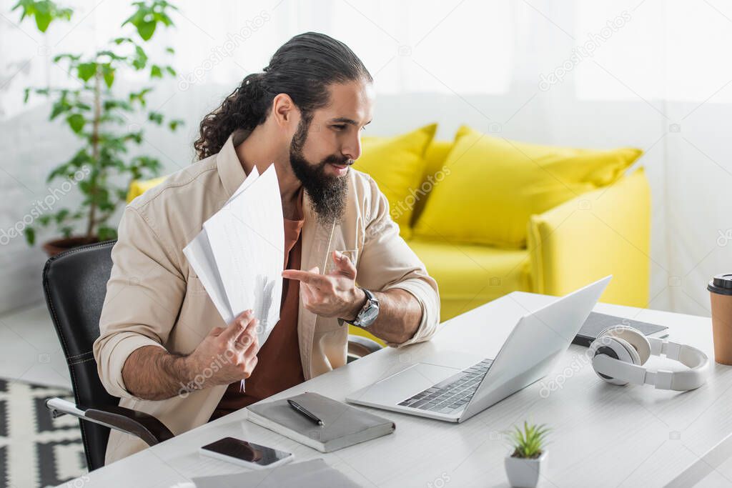 bearded hispanic freelancer pointing at documents during video call on laptop at home
