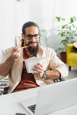 Evde öğle yemeği için Çin eriştesi yerken dizüstü bilgisayara bakan sakallı Latin serbest yazar.