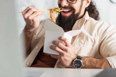Kol saatindeki sakallı adamın ön planda Çin eriştesi yerken kısmi görüntüsü.