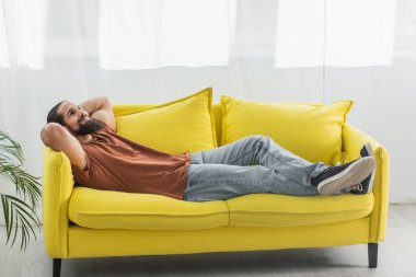 Sarı kanepede elleri başının arkasında yatarken gülümseyen sakallı İspanyol adamın tam boy görüntüsü.