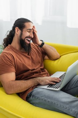 excited hispanic man laughing while using laptop on couch at home clipart