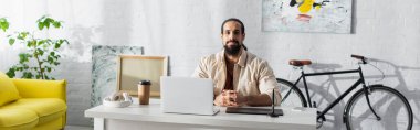 bearded latin freelancer sitting near laptop and graphic tablet in home studio, banner clipart