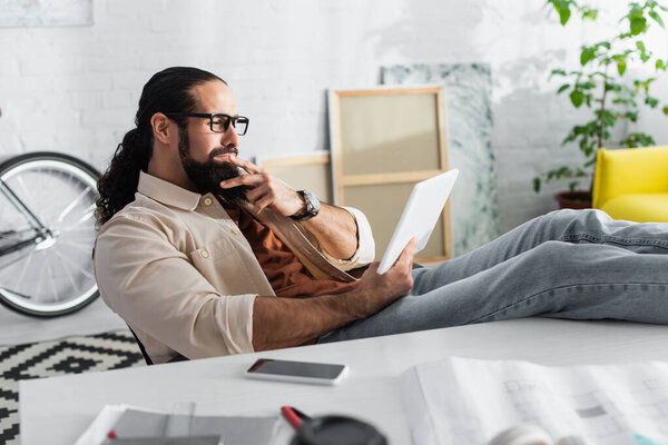 вдумчивый латинский дизайнер смотрит на цифровой планшет, сидя с ногами на столе рядом с размытым смартфоном