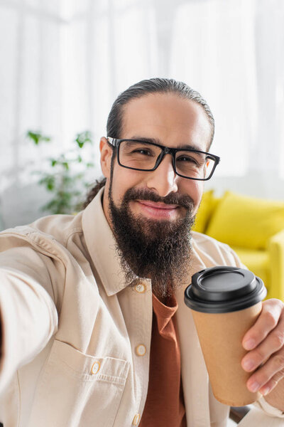 bearded hispanic man in eyeglasses smiling at camera while holding paper cup
