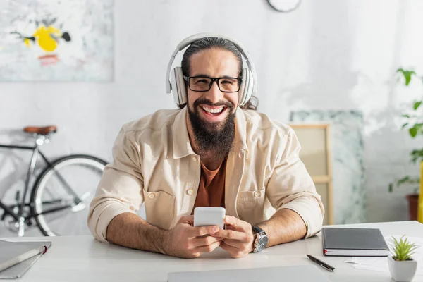 excited hispanic freelancer in headphones laughing at camera while listening podcast at home