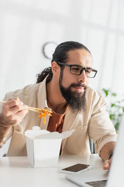 latin freelancer in eyeglasses looking at blurred laptop while eating chinese noodles