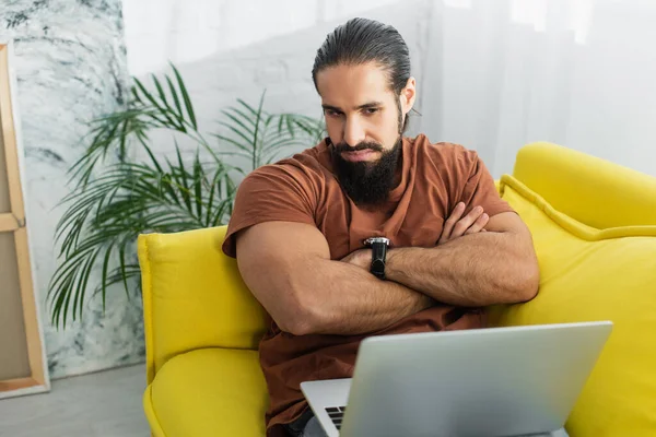 Grave Hispânico Homem Olhando Para Laptop Enquanto Sentado Sofá Casa — Fotografia de Stock