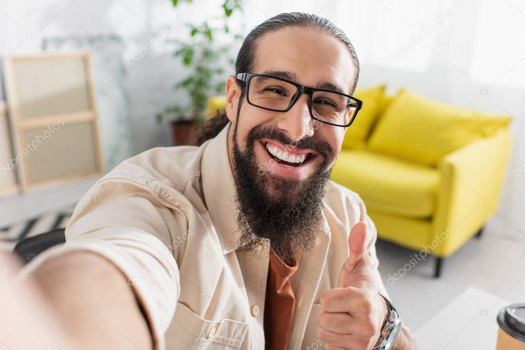 cheerful latin man in eyeglasses looking ta camera and showing thumb up on blurred foreground