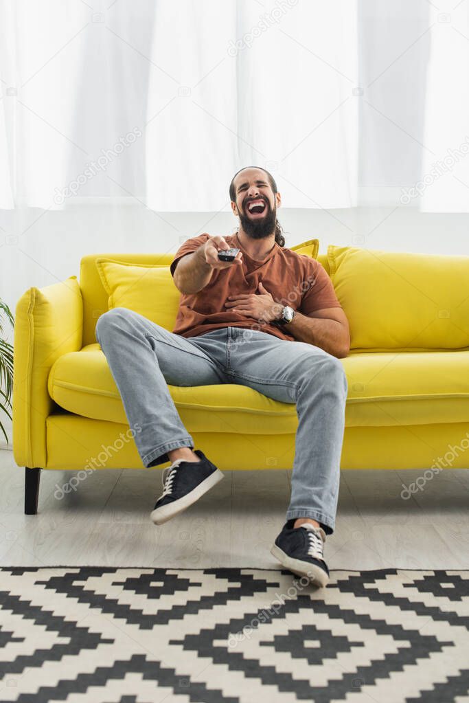 excited latin man with remote controller laughing on yellow sofa while watching tv