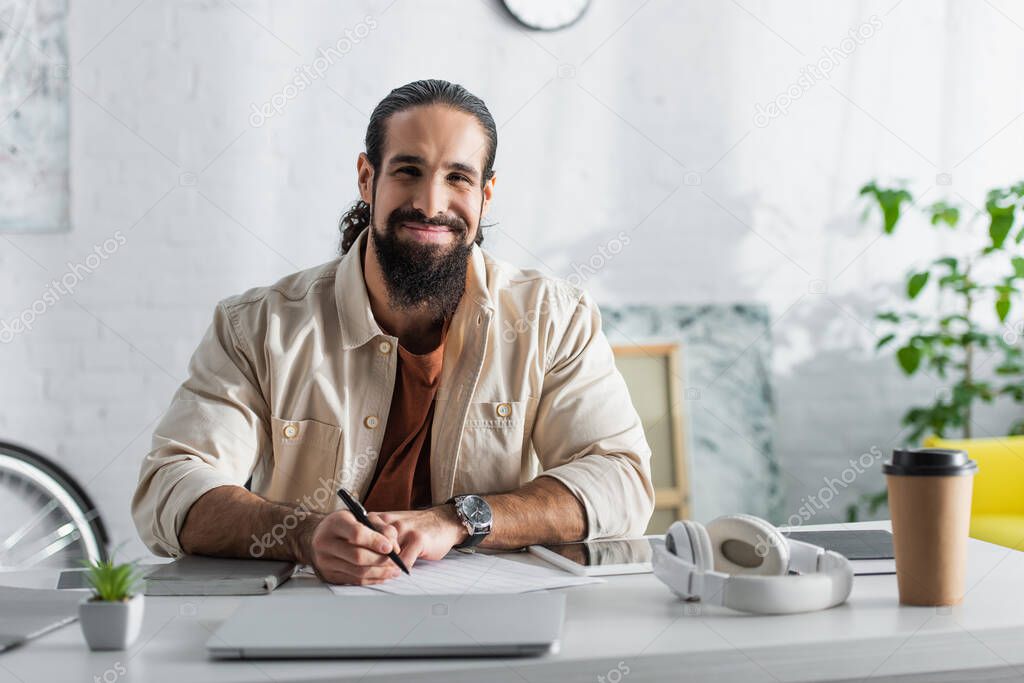 hispanic freelancer smiling at camera while working with documents near gadgets at home