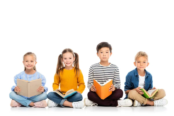 Positive multiethnische Kinder, die Bücher halten, während sie auf weißem Hintergrund sitzen — Stockfoto