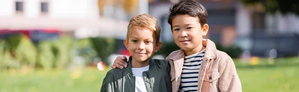 Asiatischer Junge umarmt Freund, während er Kamera im Park anschaut, Banner — Stockfoto