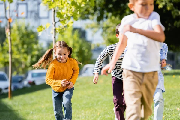 Fille souriante courant près d'amis sur le premier plan flou dans le parc — Photo de stock