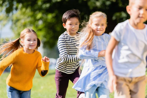 Lächelnde multiethnische Kinder, die in der Nähe von Freunden auf verschwommenem Vordergrund im Freien laufen — Stockfoto