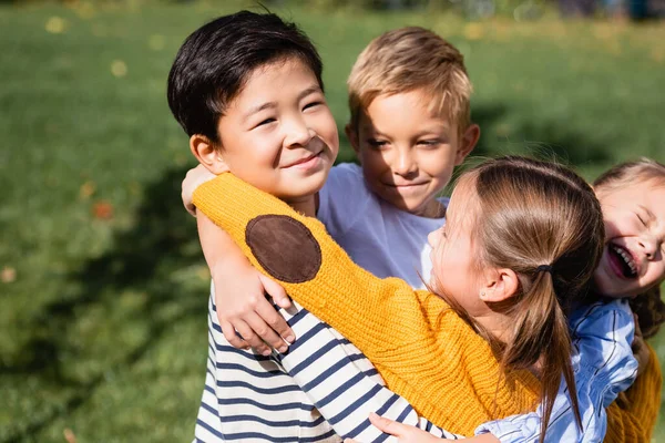 Asiático menino olhando para a câmera enquanto abraçando amigos sorridentes ao ar livre — Fotografia de Stock