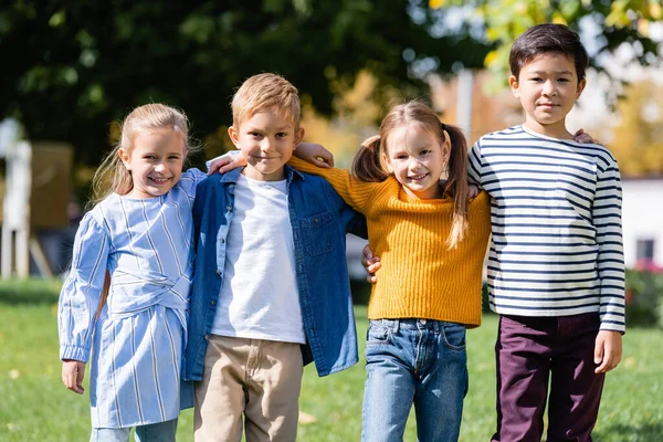 Niños multiculturales sonriendo a la cámara mientras se abrazan en el parque — Stock Photo