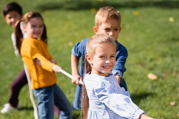 Ragazza sorridente guardando la fotocamera mentre gioca tiro alla fune con gli amici su sfondo sfocato nel parco — Foto stock