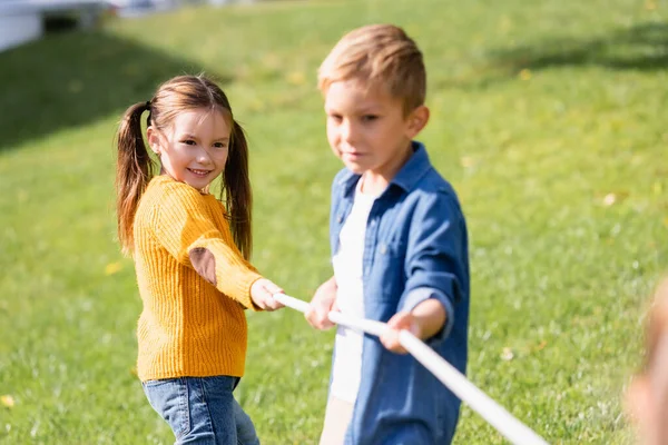 Positive child playing tug of war near friend on blurred foreground in park — Stock Photo