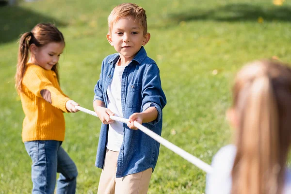 Niño sosteniendo la cuerda mientras juega tirón de la guerra con amigos en primer plano borroso en el parque - foto de stock