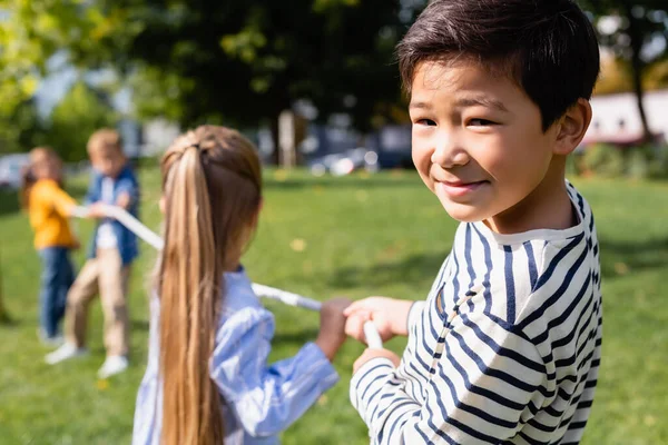 Улыбающийся азиатский мальчик смотрит в камеру, играя в перетягивание каната с друзьями на размытом фоне в парке — стоковое фото
