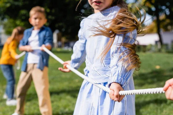 Chica sosteniendo la cuerda mientras juega tirón de la guerra con amigos en el fondo borroso en el parque - foto de stock