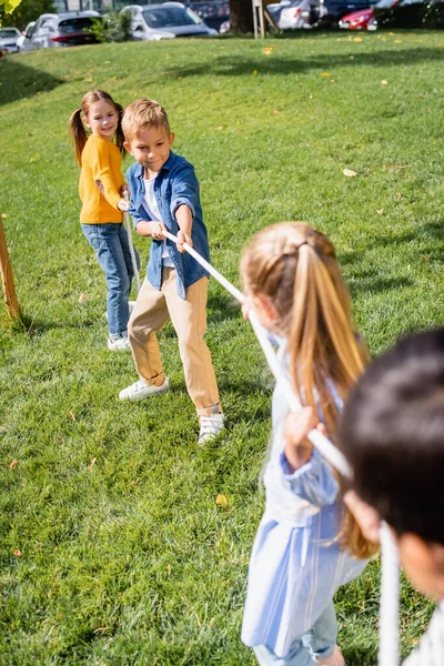 Cheerful kids plying tug of war with friends on blurred foreground in park — Stock Photo