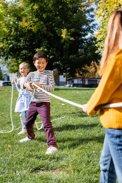 Sorridente asiatico ragazzo tirando corda mentre gioca rimorchiatore di guerra con gli amici in primo piano offuscata nel parco — Foto stock